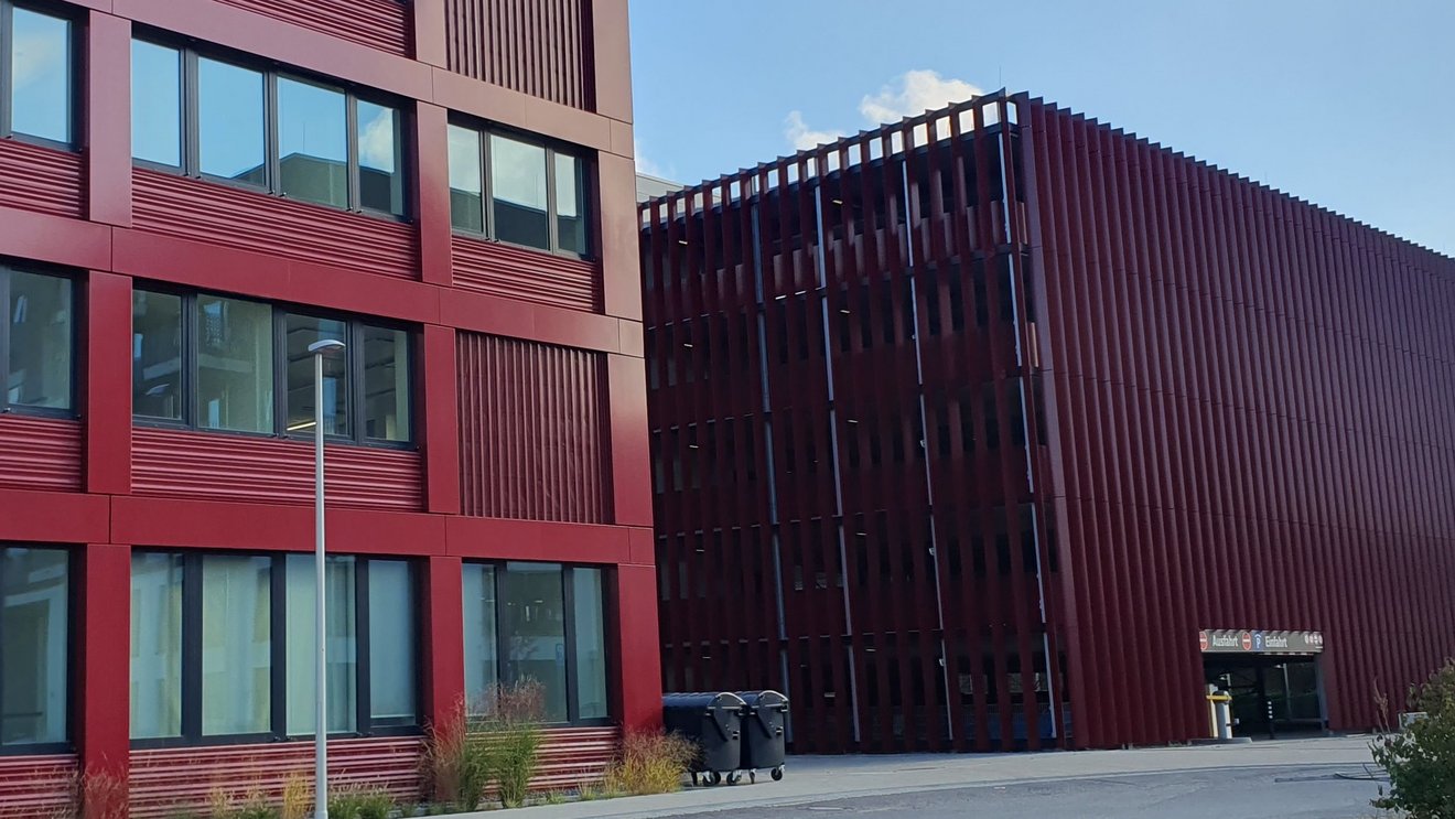 Dienstgeb&auml;ude des ZFB in Bonn mit roter Fassade und gro&szlig;en Fenstern, rechts daneben Parkhaus-Einfahrt Kachel Vorschau